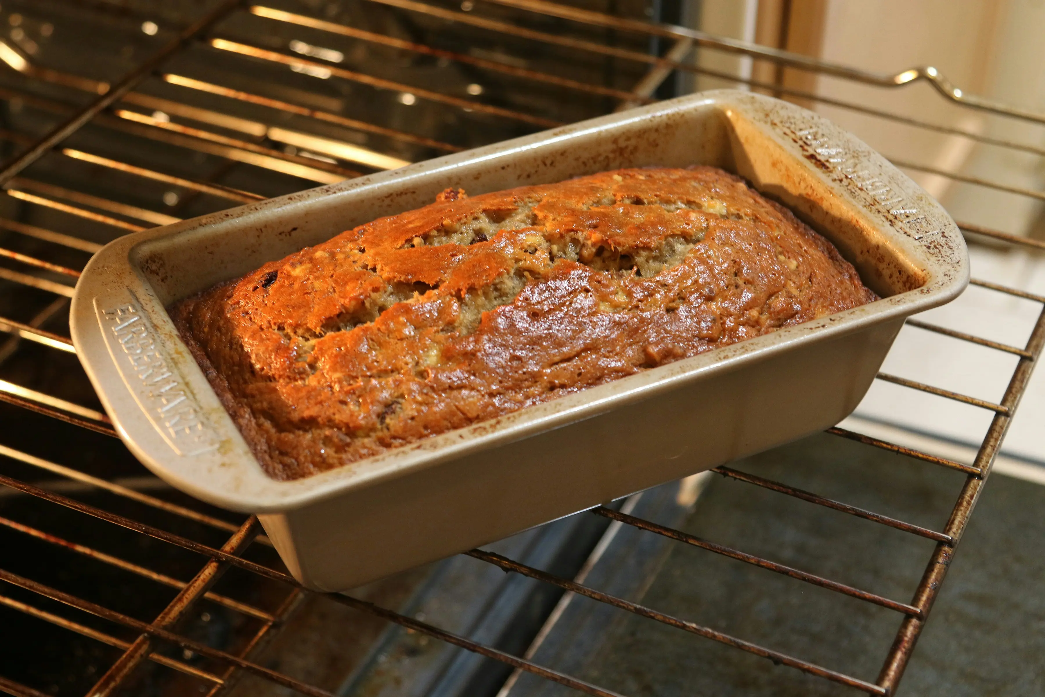 Banana bread loaf in a gold loaf pan inside the oven, golden brown crust with a natural centre crack, baking on oven rack