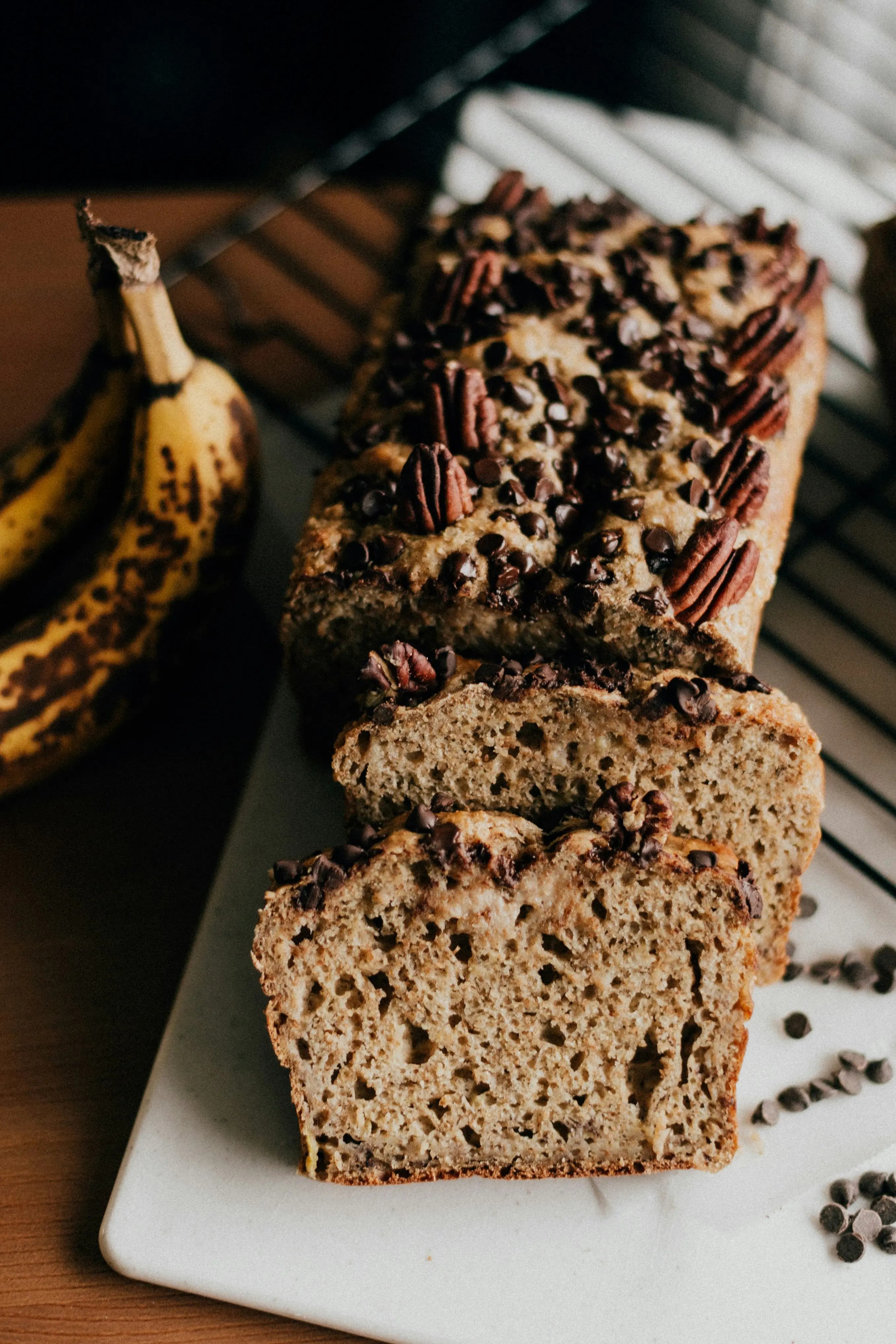 Banana bread loaf with chocolate chips and pecan halves on top, sliced to show moist crumb, overripe bananas in background, moody natural light
