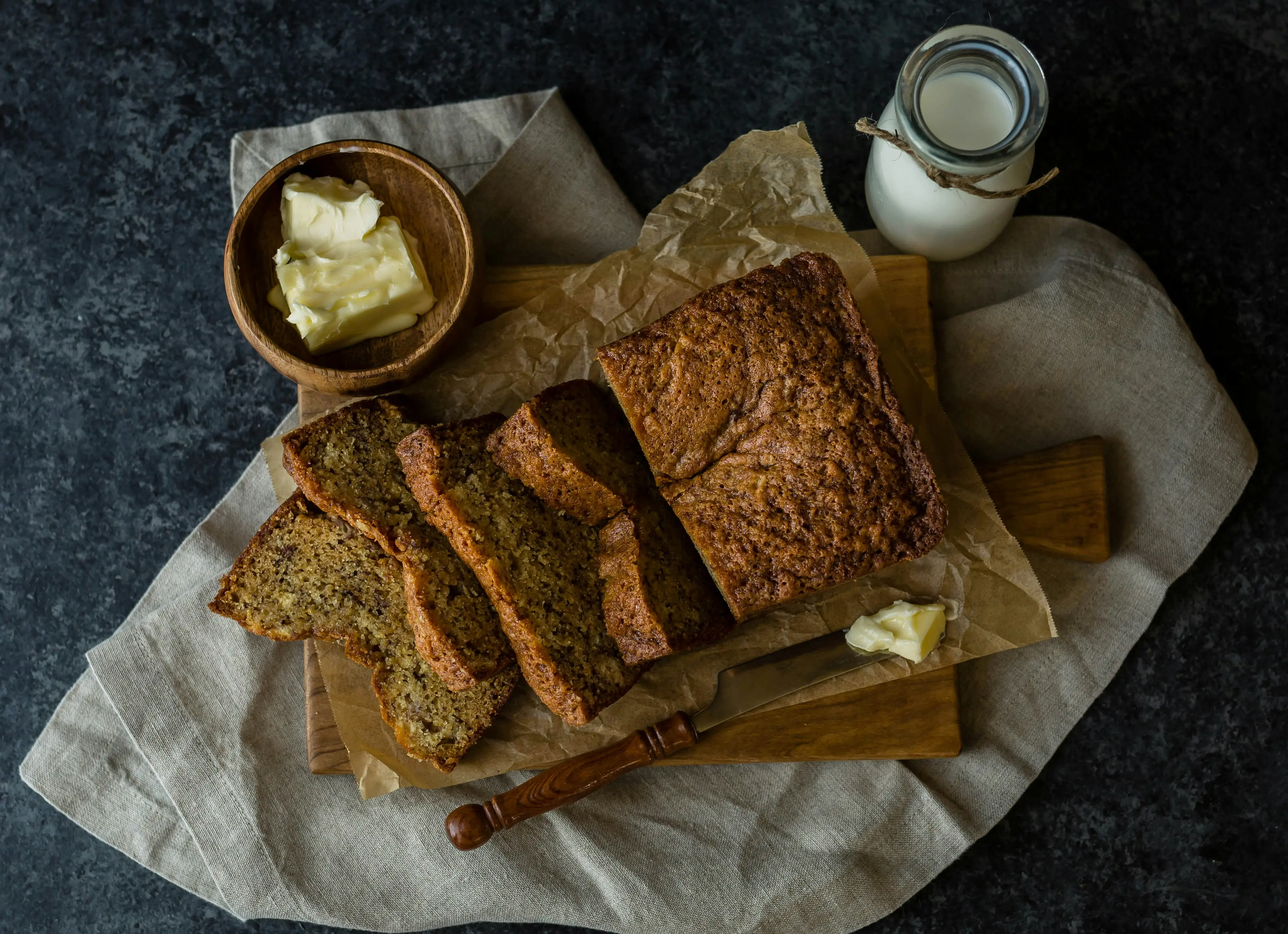 Sliced banana bread loaf on a wooden board with parchment paper, butter and milk jar beside it, overhead moody food photography
