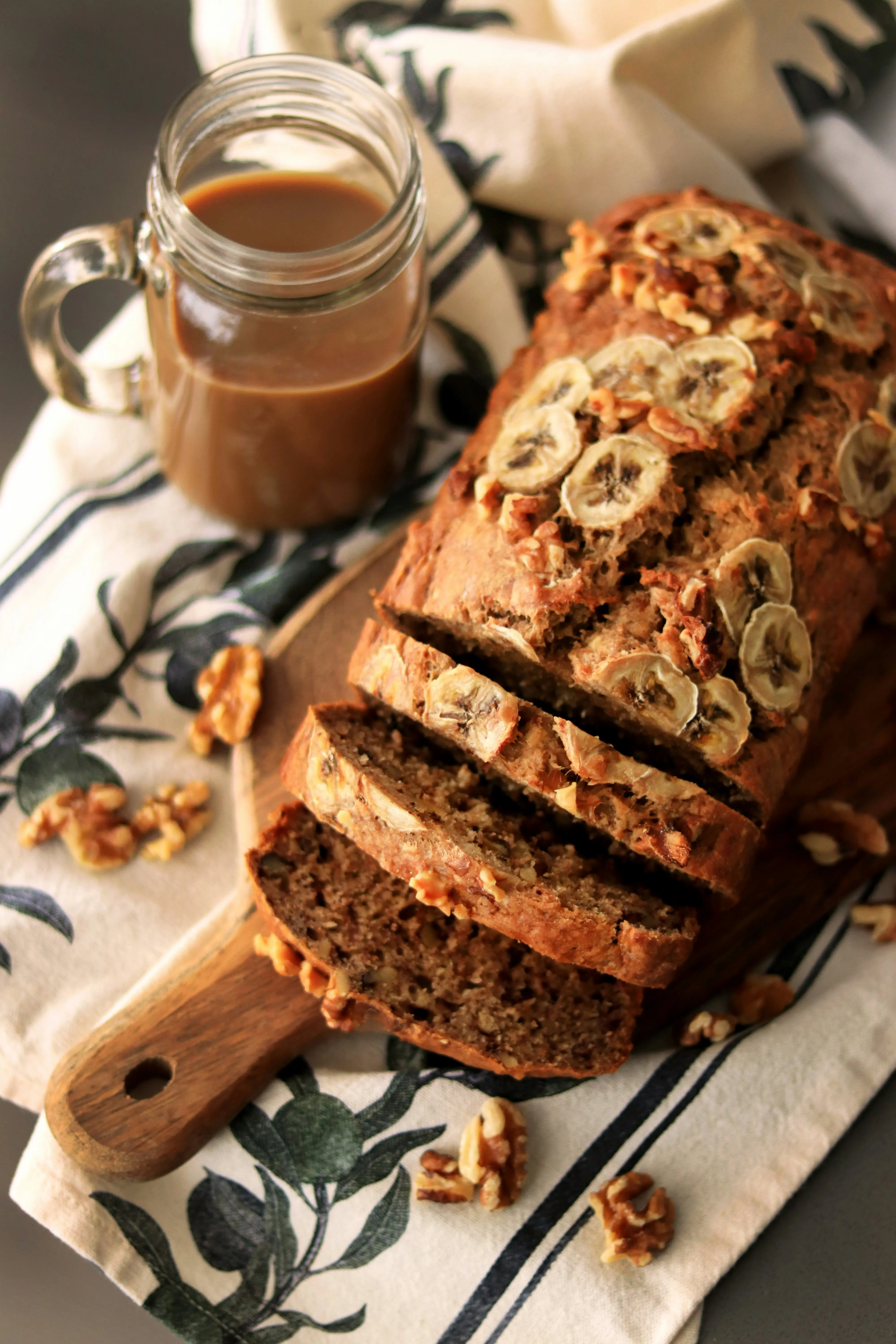 Sliced banana walnut bread on a wooden cutting board with dried banana slices on top, walnuts scattered around, jar of caramel in background