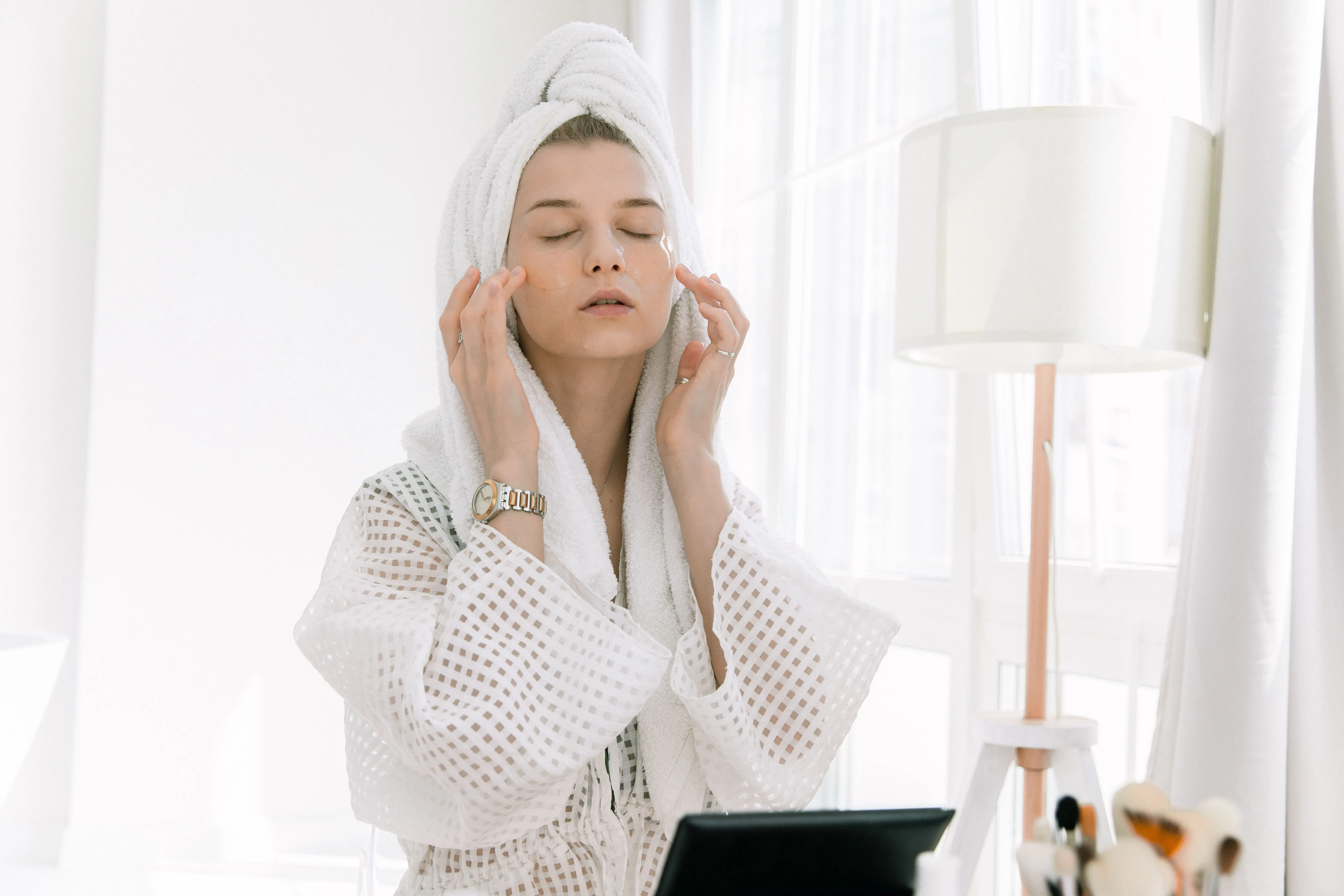 Morning skincare ritual for glass skin — woman applying eye patches in white robe natural window light